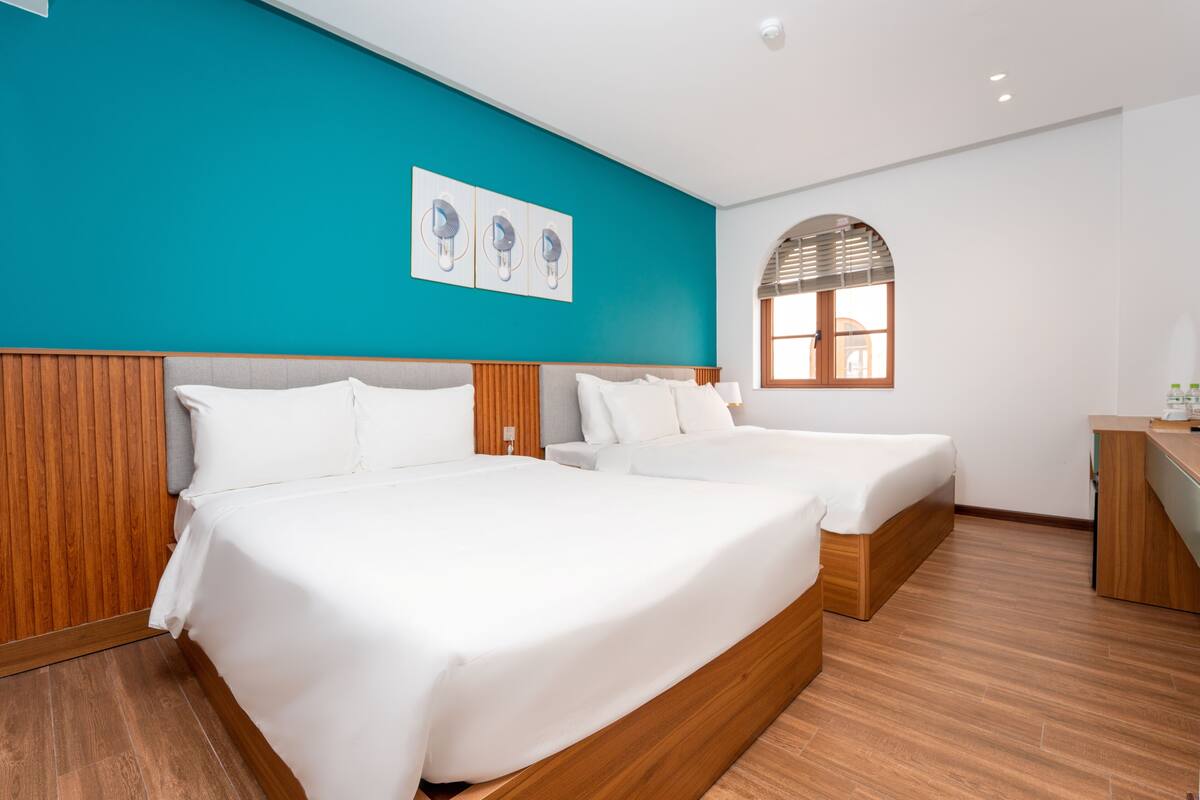 A hotel room with two beds, wooden headboards, a teal accent wall, and a window with blinds.