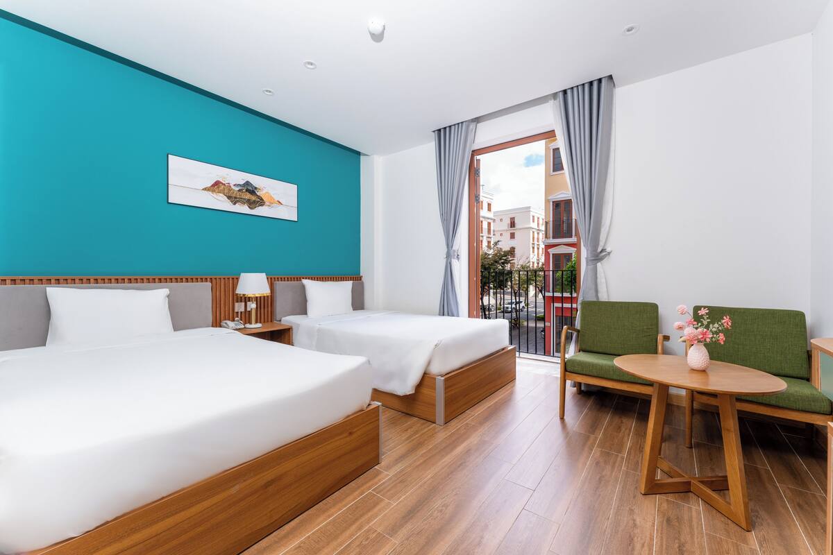 A hotel room with two beds, a green sofa, a small table, and a balcony with a view of buildings.