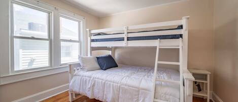 Una cama individual superpuesta en una habitación con ventanas amplias, una pared blanca y suelo de madera.