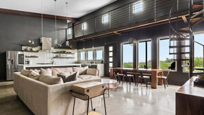 A spacious living area with a sofa, dining table, and chairs, a kitchen with modern appliances, and large windows offering a view. - Enchanted Rock Ridge / Cowboy Pool / Hot Tub / Firepit (Fredericksburg)