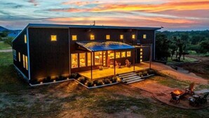 A modern two-story house with a large glass front, a covered porch, and a fire pit area. - Enchanted Rock Ridge / Cowboy Pool / Hot Tub / Firepit (Fredericksburg)