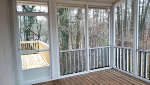A covered porch with a wooden deck and a railing, overlooking a forested area.