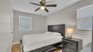 A bedroom with a bed, a nightstand, a lamp, a ceiling fan, and two windows with blinds.