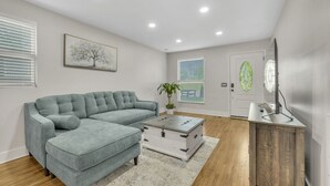 A living room with a sectional sofa, a coffee table, a potted plant, a door with a glass panel, and a television on a wooden stand.