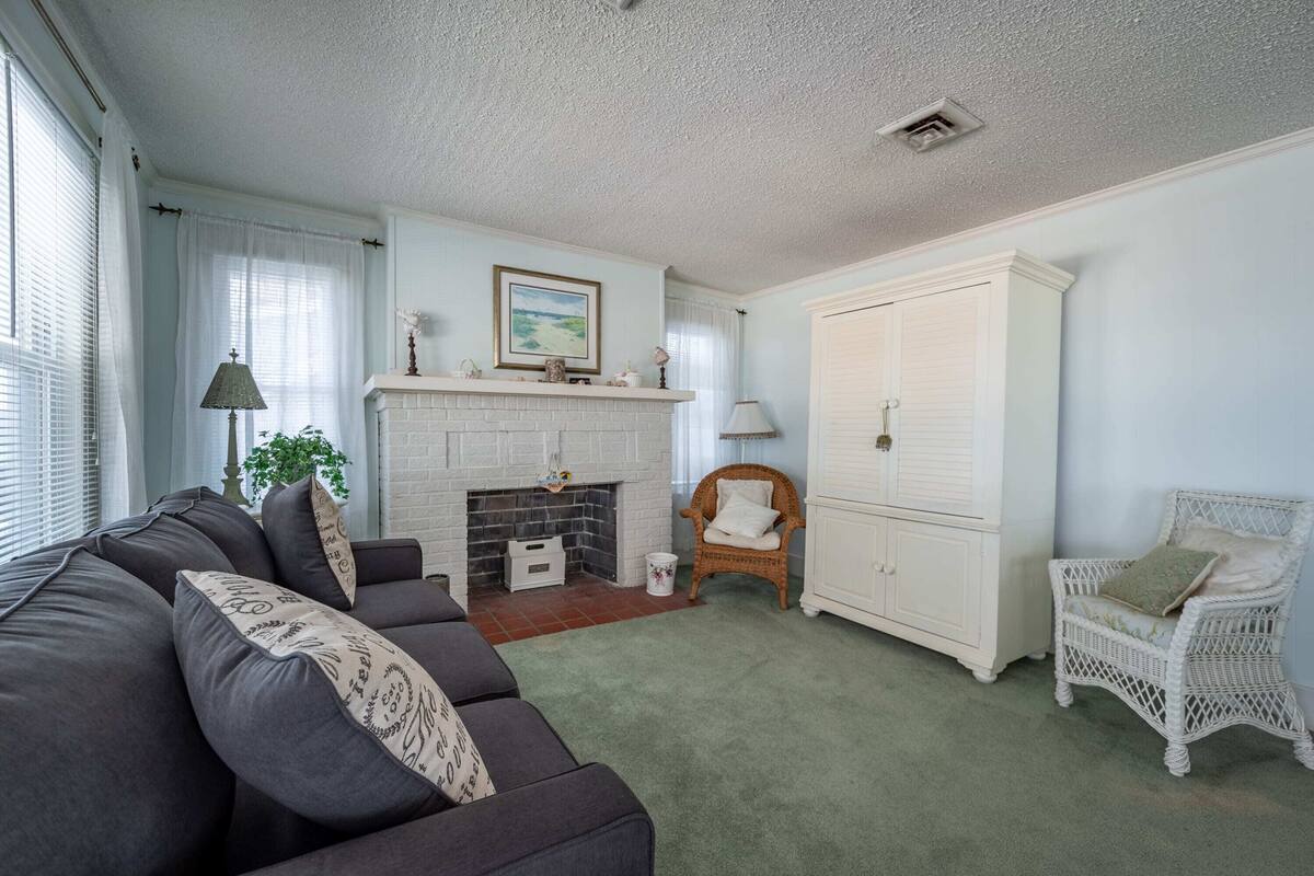 Un salon avec une cheminée, une armoire blanche, un fauteuil en osier et un canapé de couleur foncée.