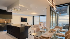 A modern kitchen with a central island, a dining area with a glass table, and a view of the sky through large windows. - Villa Luxe by Atlantic Holiday (Ribeira Brava)
