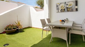 Une terrasse avec une table et des chaises, entourée de plantes en pot et ornée d’un tableau accroché au mur.