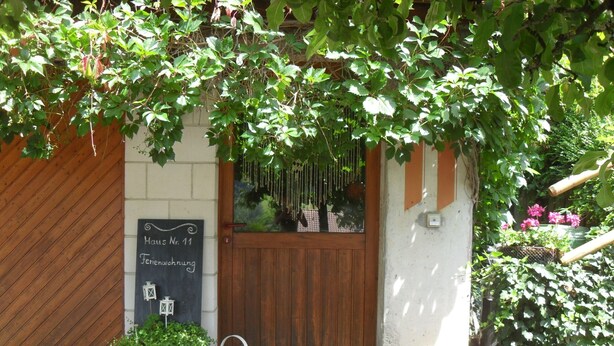 Une porte en bois avec une enseigne au-dessus, un pot de fleurs et un arrosoir posés sur le sol.