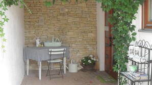 Un mur de pierres, une table recouverte d’une nappe à carreaux, une chaise, un panier et une étagère en métal avec divers objets.