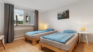 A room with two wooden beds, a window with curtains, a radiator, a small table with a lamp, and a painting on the wall. - Apartment 'Fewo Max' with Shared Terrace, Balcony and Wi-Fi (Ettenheim Münchweier)