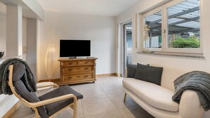 A living room with a sofa, a chair, a TV, a wooden dresser, and a window with a view. - Apartment 'Fewo Max' with Shared Terrace, Balcony and Wi-Fi (Ettenheim Münchweier)