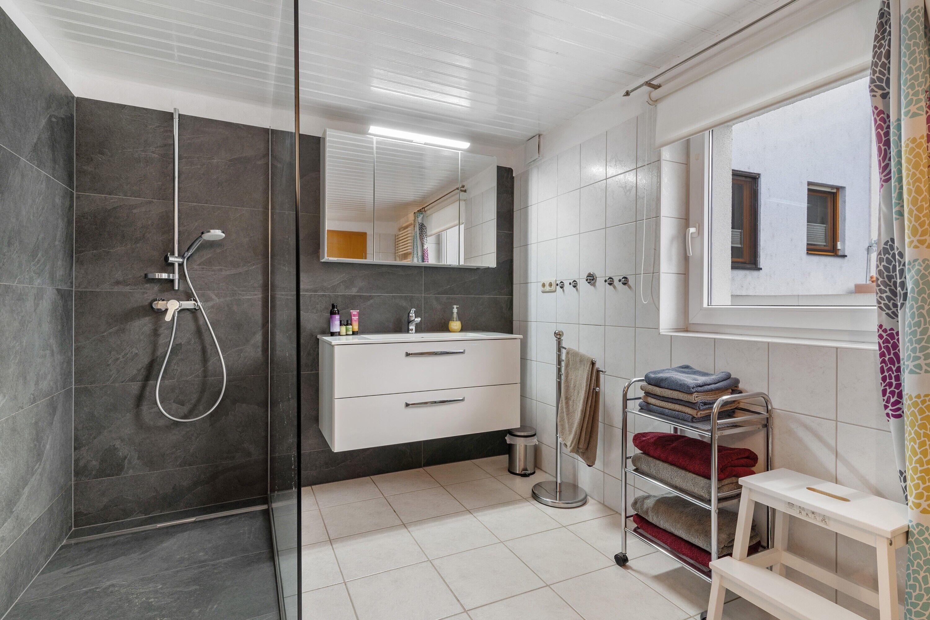 Un baño moderno con cabina de ducha de vidrio, tocador blanco, espejo, perchero y ventana con vistas a edificios.