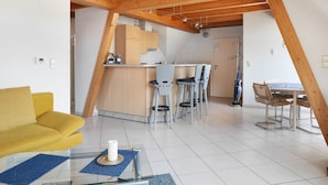 Eine moderne Küche mit Holzbalken, ein Essbereich mit Tisch und Stühlen und ein gelbes Sofa in einem hellen Wohnraum.