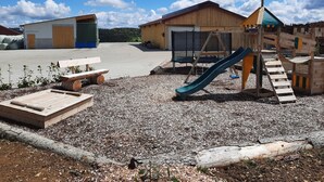 A playground with a slide and swings, surrounded by a gravel area with benches and a wooden structure.