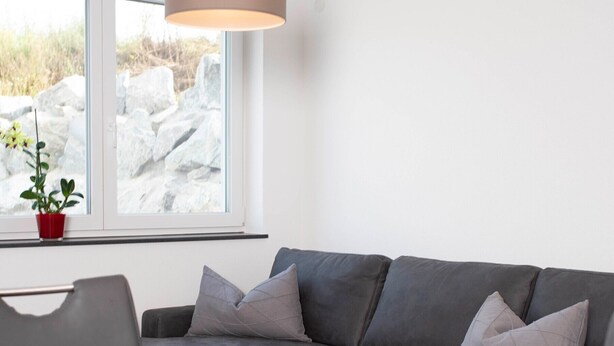 A modern living room with a grey sofa, a wooden dining table with fruit, a hanging lamp, and a window with a view of rocks and plants.