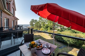 A balcony with a red umbrella, a table with a vase of flowers, and a view of the sea.