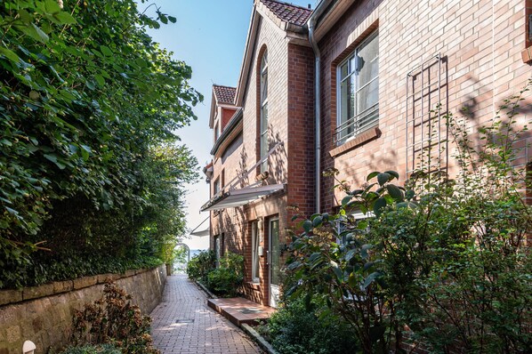 A narrow brick pathway flanked by lush greenery and a building with a traditional facade.