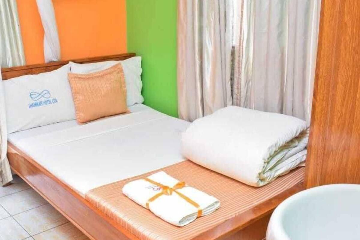 Una habitación de hotel con una cama, un almohadón, una manta y una toalla sobre un banco de madera.