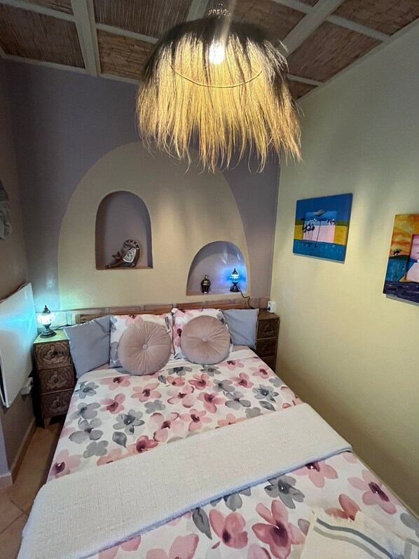 Una camera da letto con una coperta a fiori, due comodini, una lampada e due mensole a parete con soprammobili.