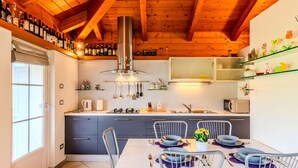 Una cocina moderna con vigas de madera, una zona de comedor con una mesa blanca y sillas metálicas, y diversos electrodomésticos.