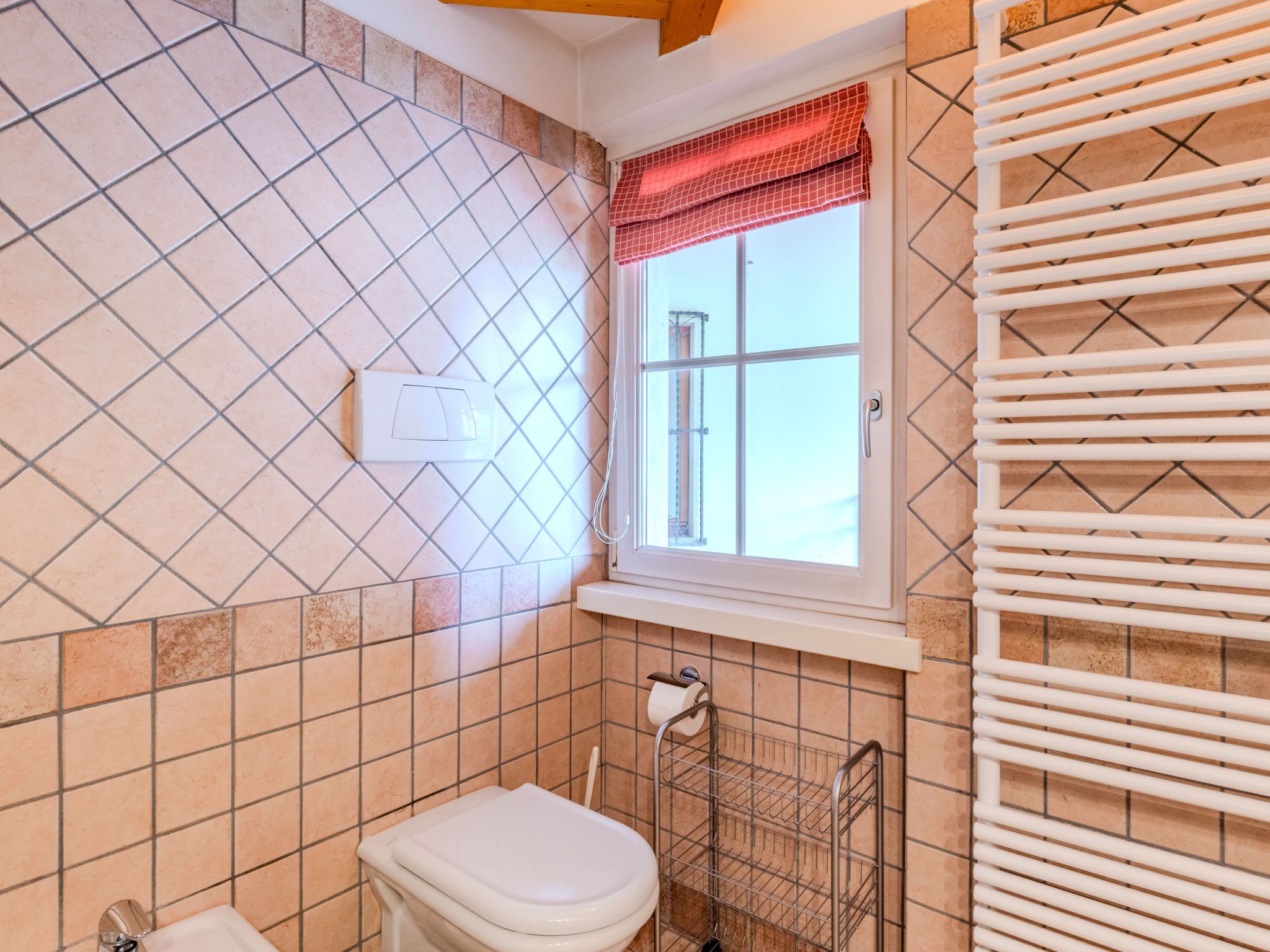 A bathroom with a toilet, a window with a red valance, a towel rack, and a shower area.