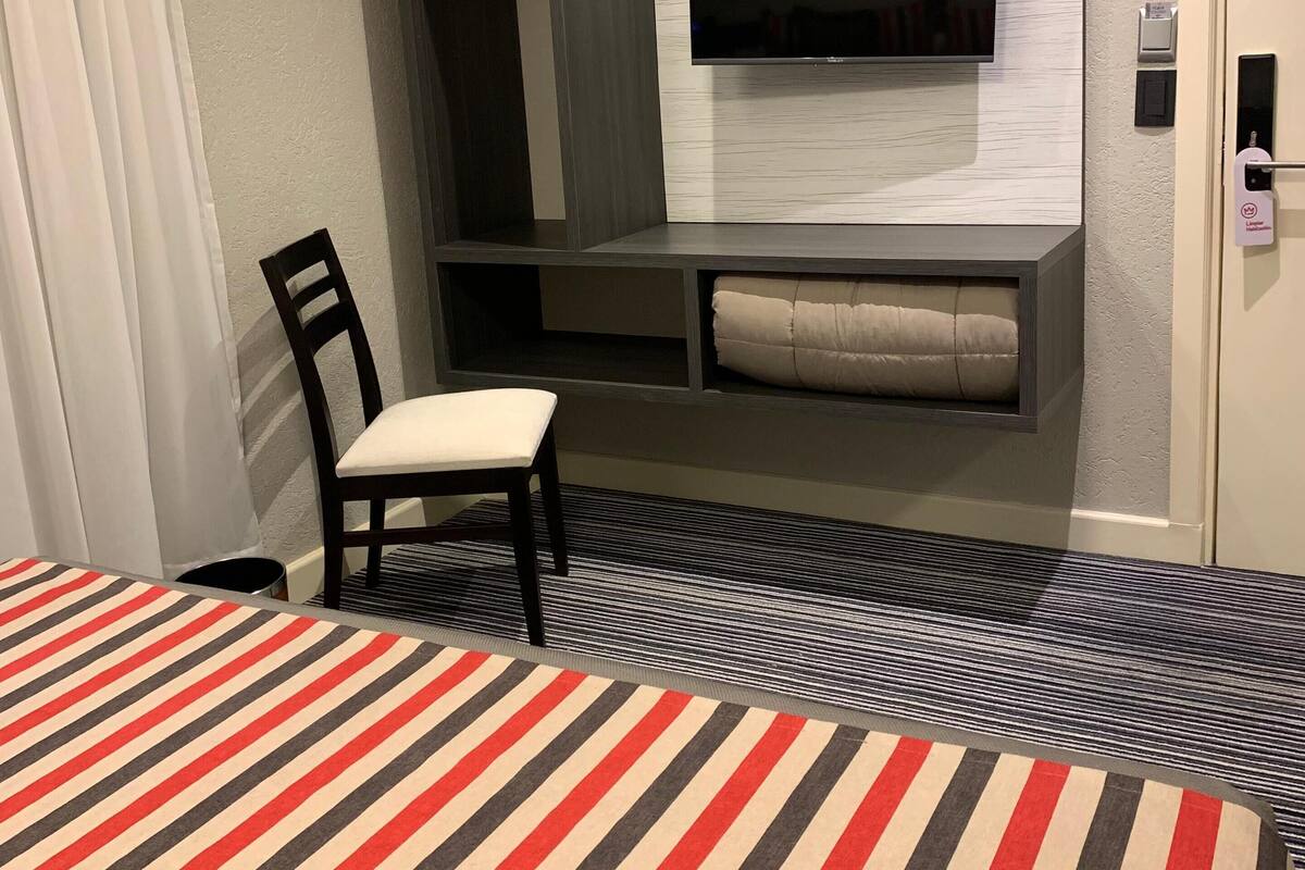 Una habitación de hotel con una cama, una silla, un televisor montado en la pared, un armario y una mesita.