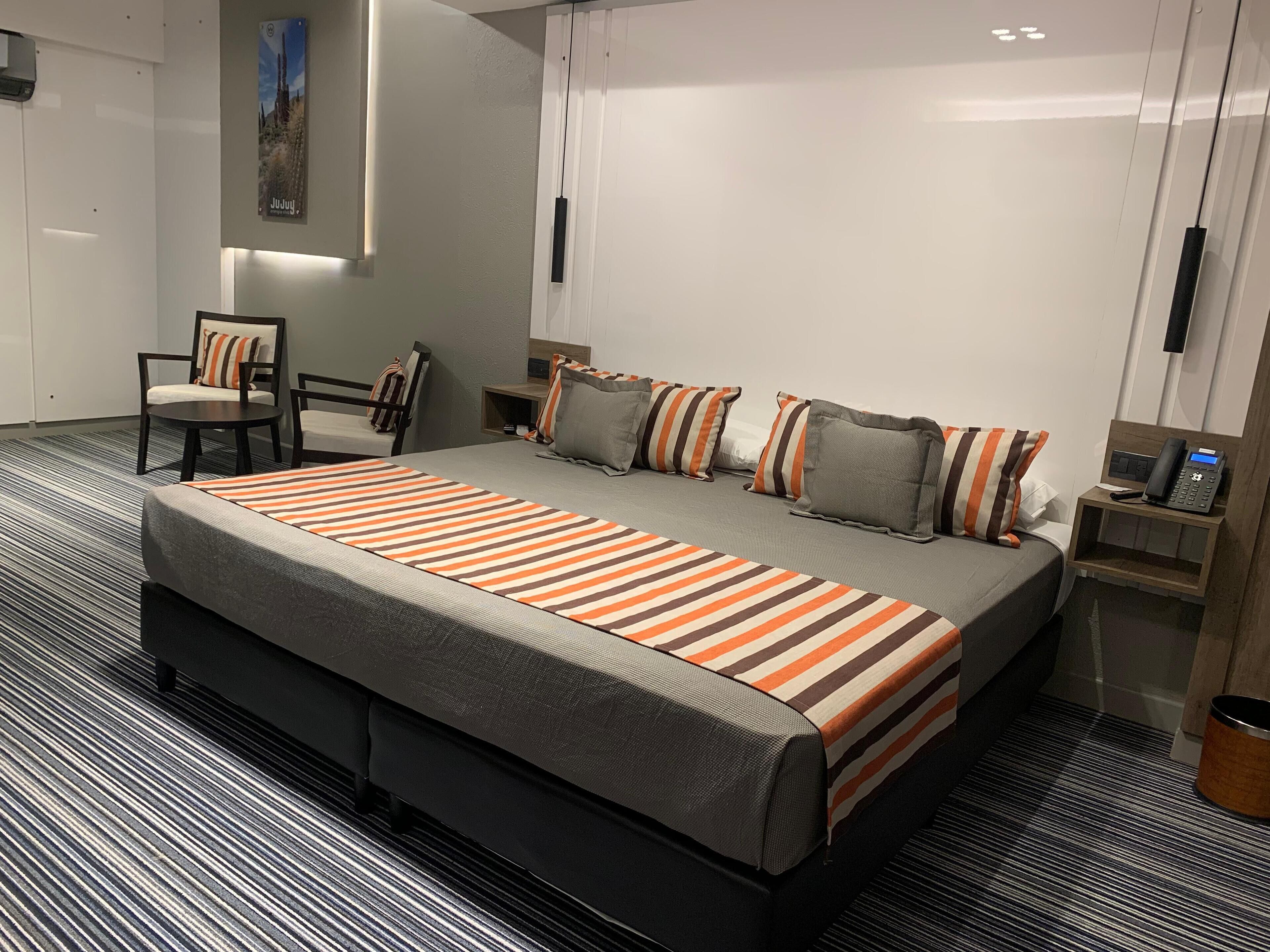 A modern hotel room with a large bed, a chair, a bedside table with a phone, and a small cabinet.