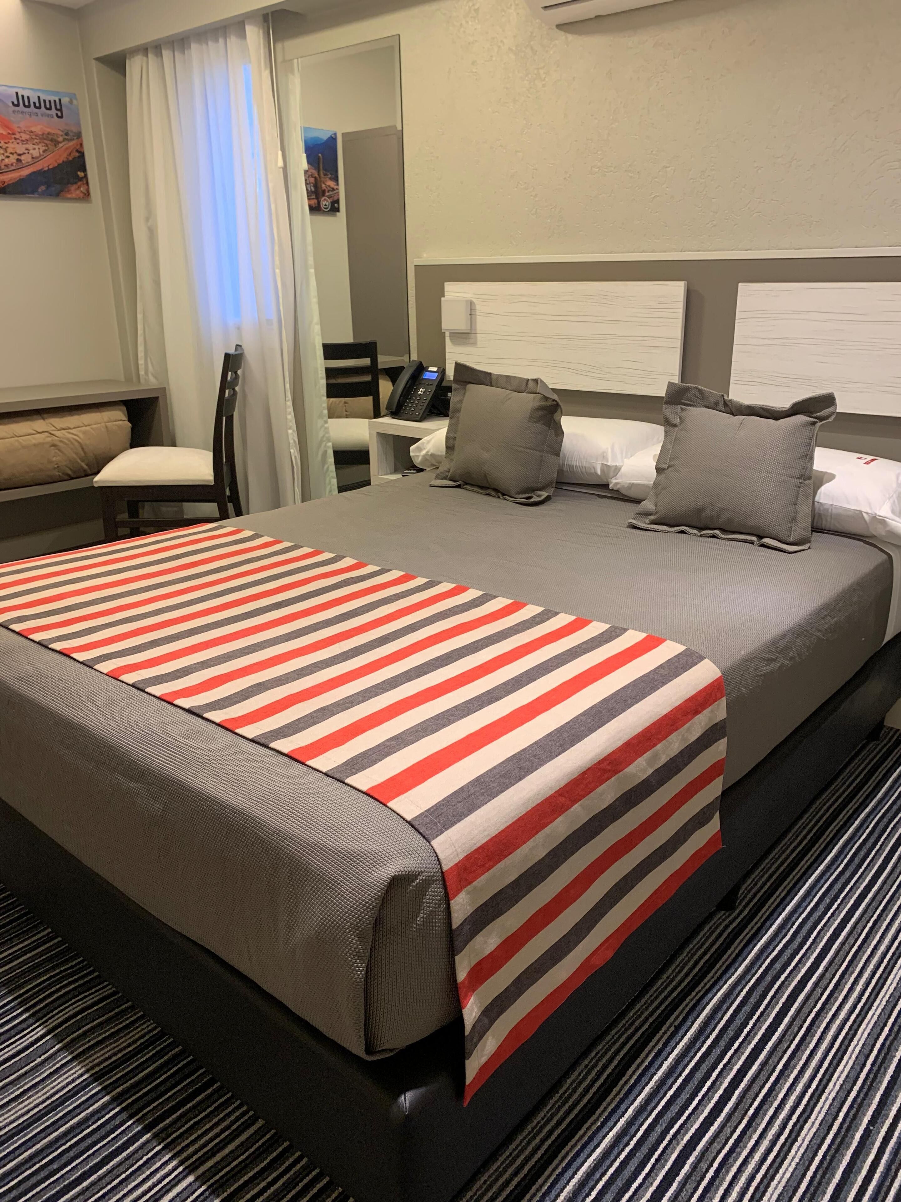 Habitación de hotel con una cama grande, un escritorio con teléfono, una silla y vistas al exterior por una ventana.