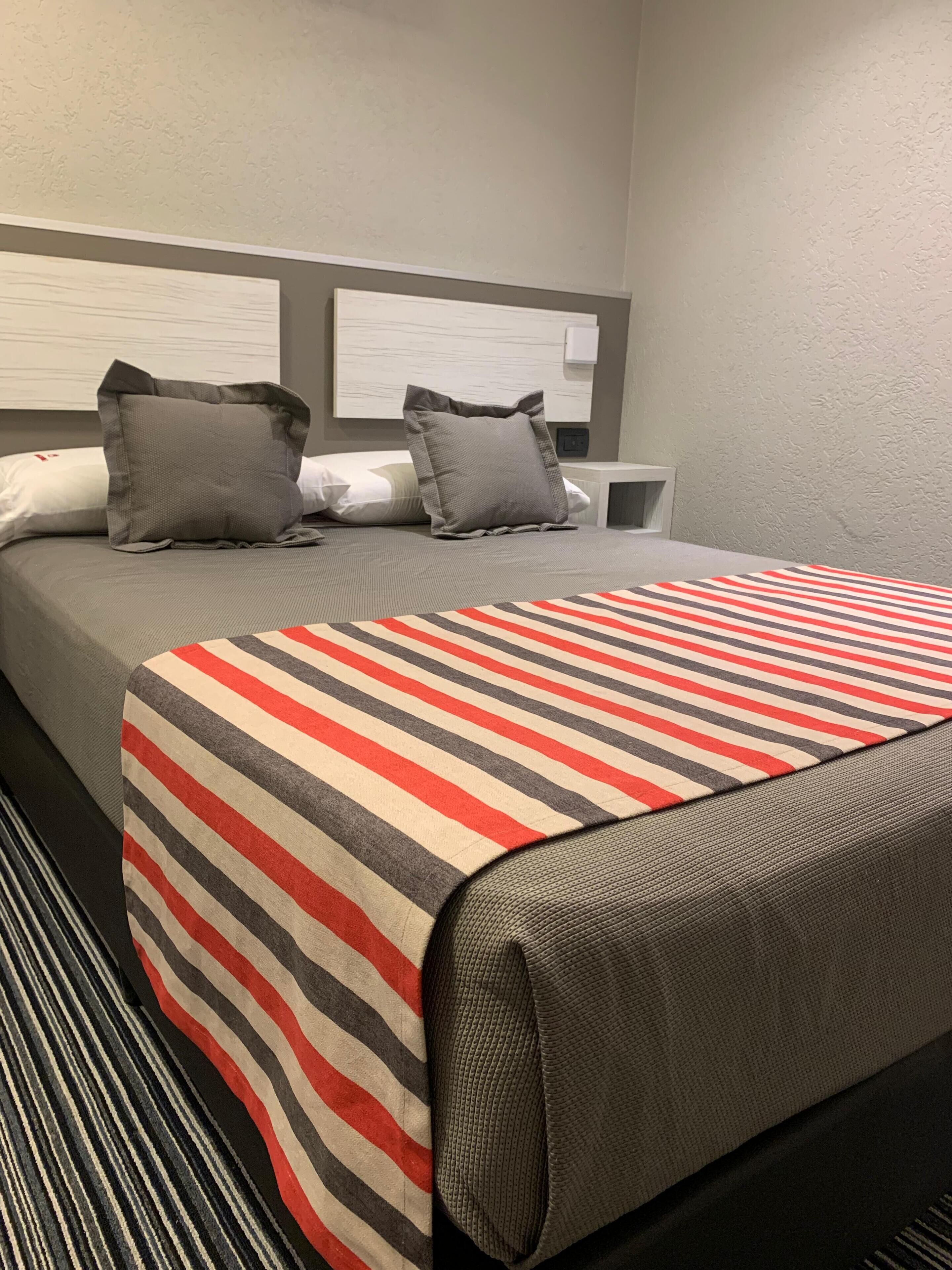 A hotel room with a bed, two pillows, a bedside table, and a wall-mounted light.