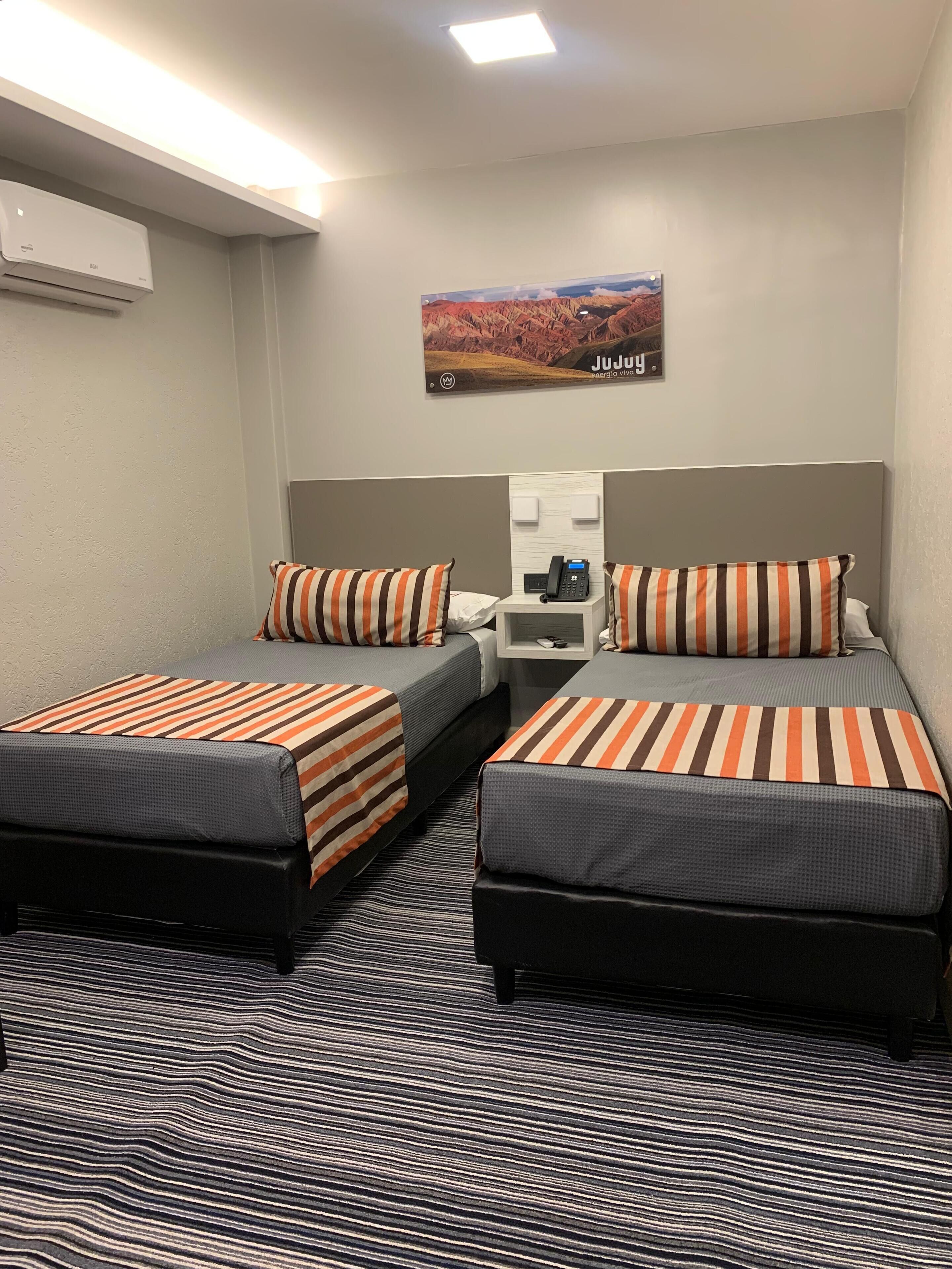 Een hotelkamer met twee bedden, gestreepte beddengoed, een nachtkastje met telefoon, een schilderij van een landschap en een kamerbreed tapijt met dessin.