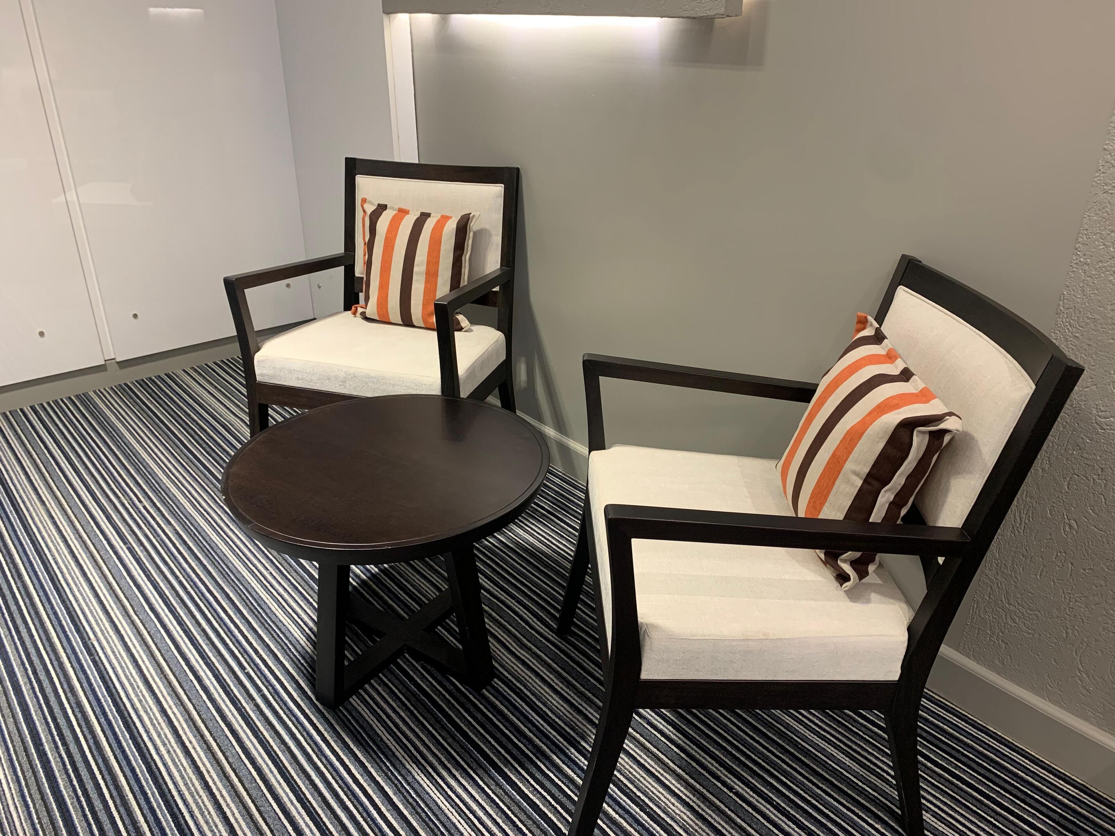 Two chairs with striped pillows, a small round table, and a striped carpet in a room with a plain wall and a light fixture.
