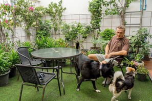 Uma pessoa sentada a uma mesa redonda, com cadeiras, rodeada por plantas em vasos e um pequeno cão.
