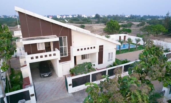 A two-story house with a balcony, a car parked in the driveway, and a swimming pool in the background.