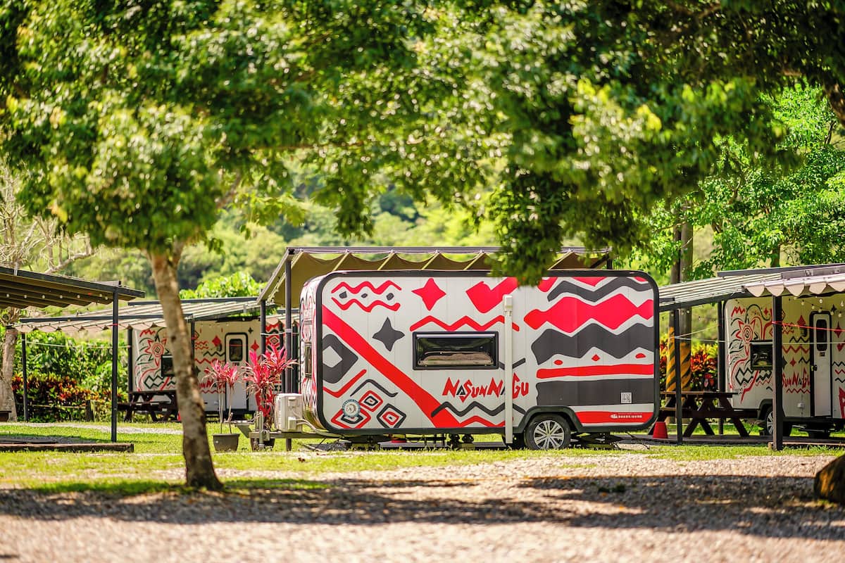A caravan with a distinctive design, parked in a park-like setting with trees and a picnic area in the background.
