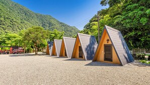 A row of triangular-roofed cabins with a gravel ground, set against a backdrop of lush green hills and clear blue sky. - NA SUN NA GU (Nan-ao)