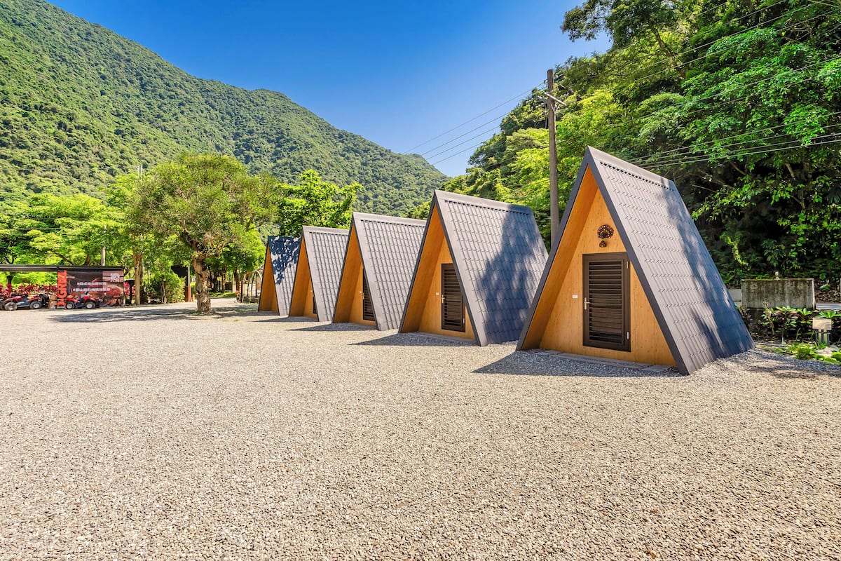 A row of triangular-roofed cabins with a gravel ground, set against a backdrop of lush green hills and clear blue sky.
