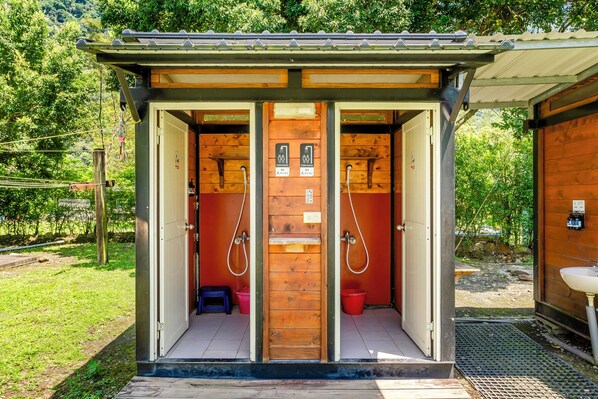 Una estructura de madera con dos puertas, cada una que da a un cubículo de baño. Hay dos cubos rojos y un taburete pequeño al lado de las puertas.