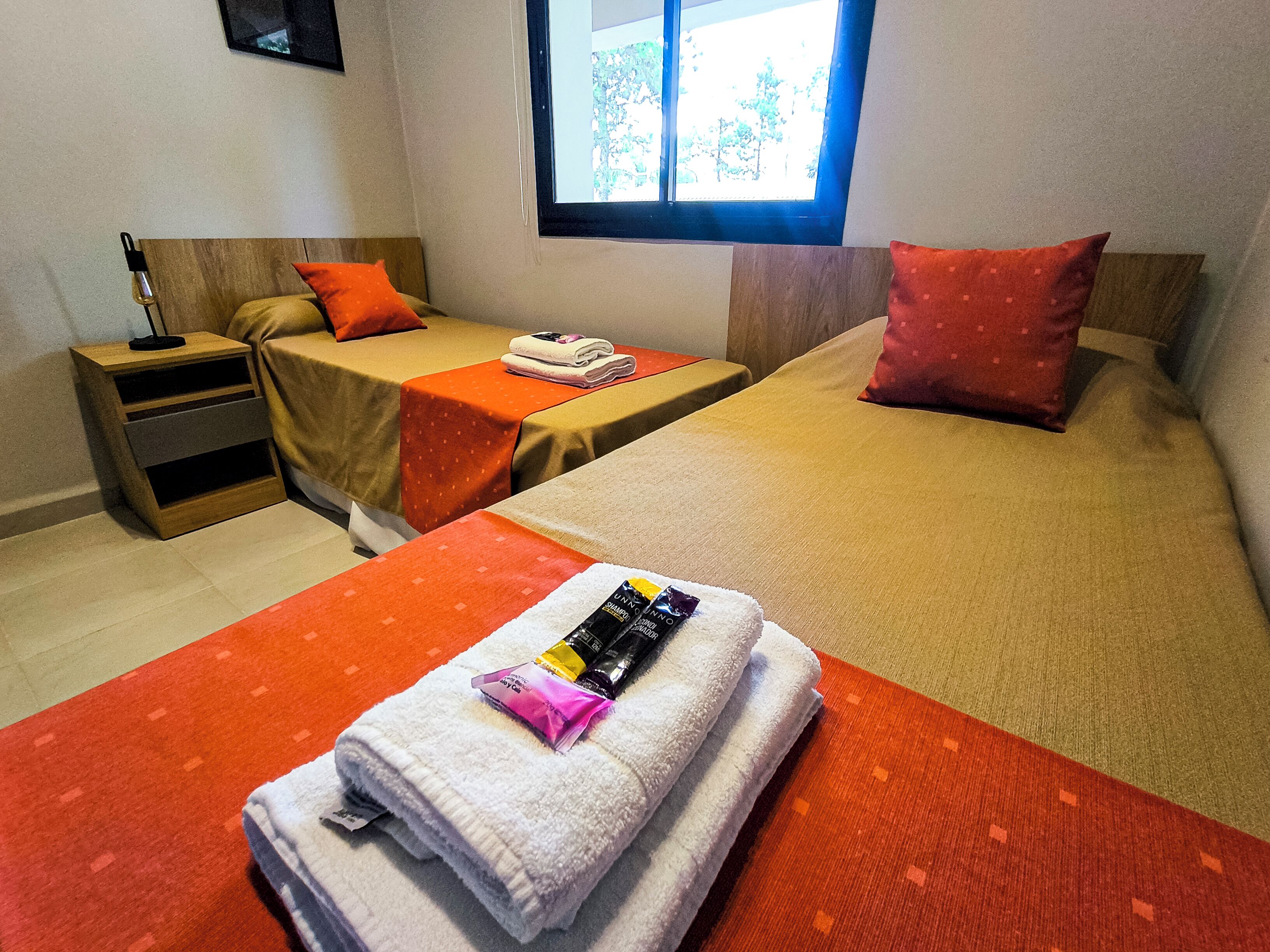 Habitació d'hotel amb dos llits, cadascun amb un coixí vermell i una tovallola blanca. Es veu una taula de nit amb una làmpada i una taula petita amb una tovallola doblegada i productes de tocador.