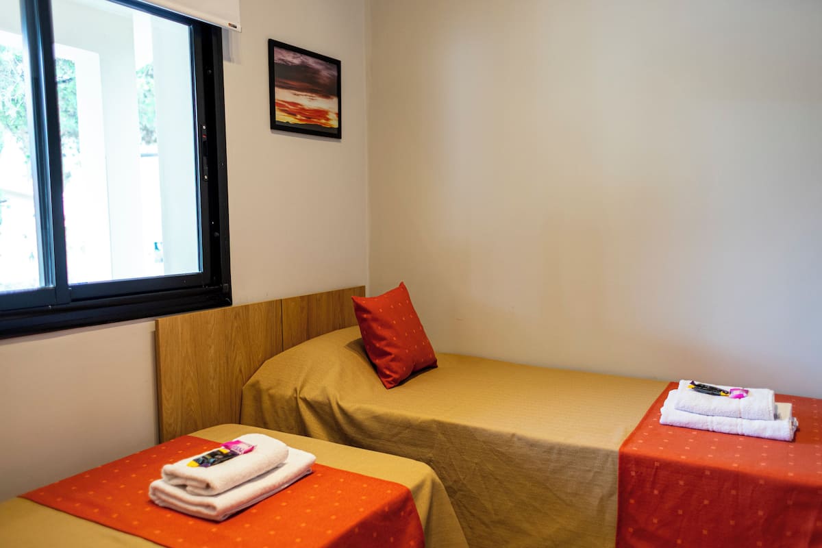 Une petite pièce avec deux lits, chacun recouvert d’une couverture rouge et beige. Il y a une fenêtre sur le côté gauche et un tableau encadré accroché au mur au-dessus des lits.