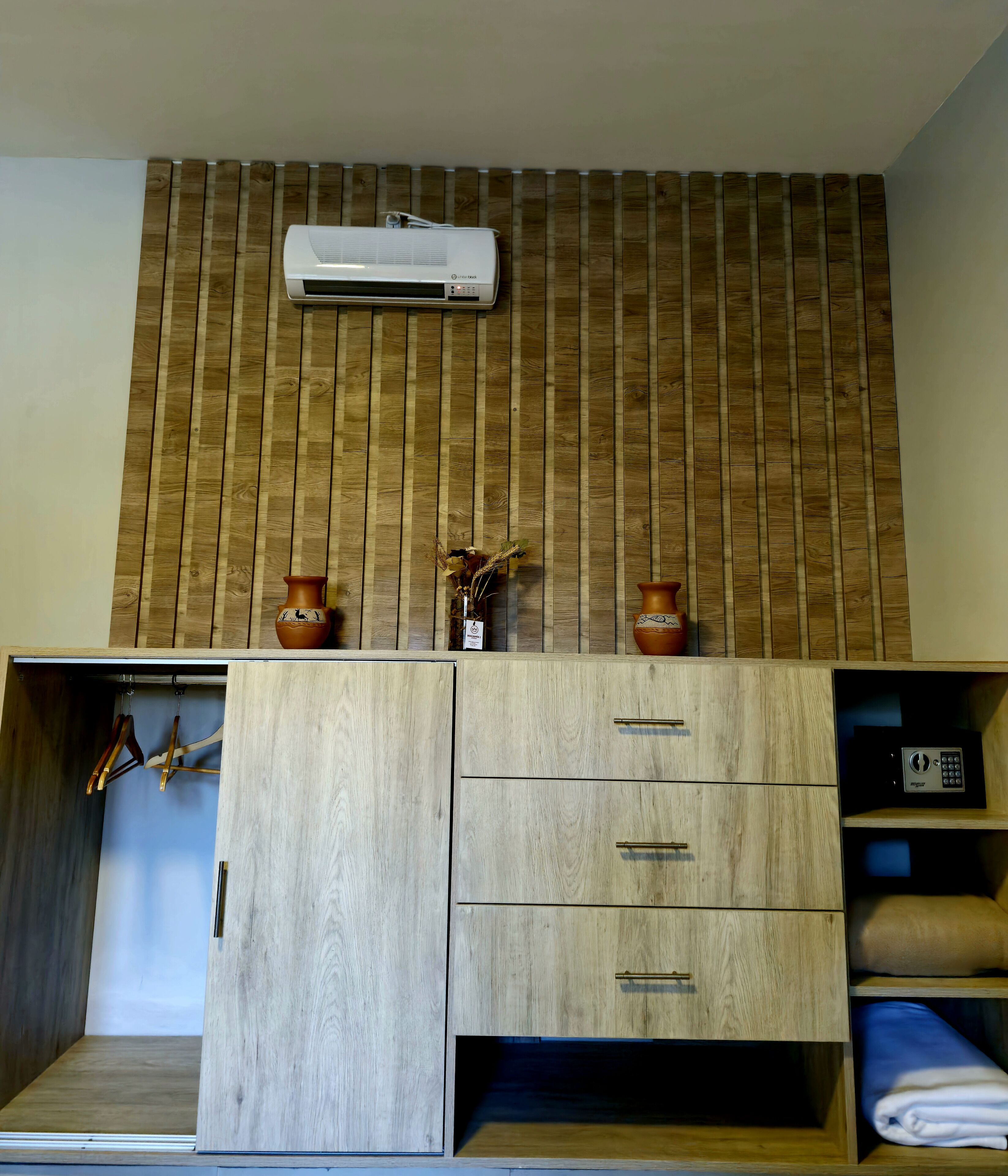 Una habitación con pared de listones de madera, aire acondicionado integrado, un mueble de madera con cajones y un estante con una manta doblada.