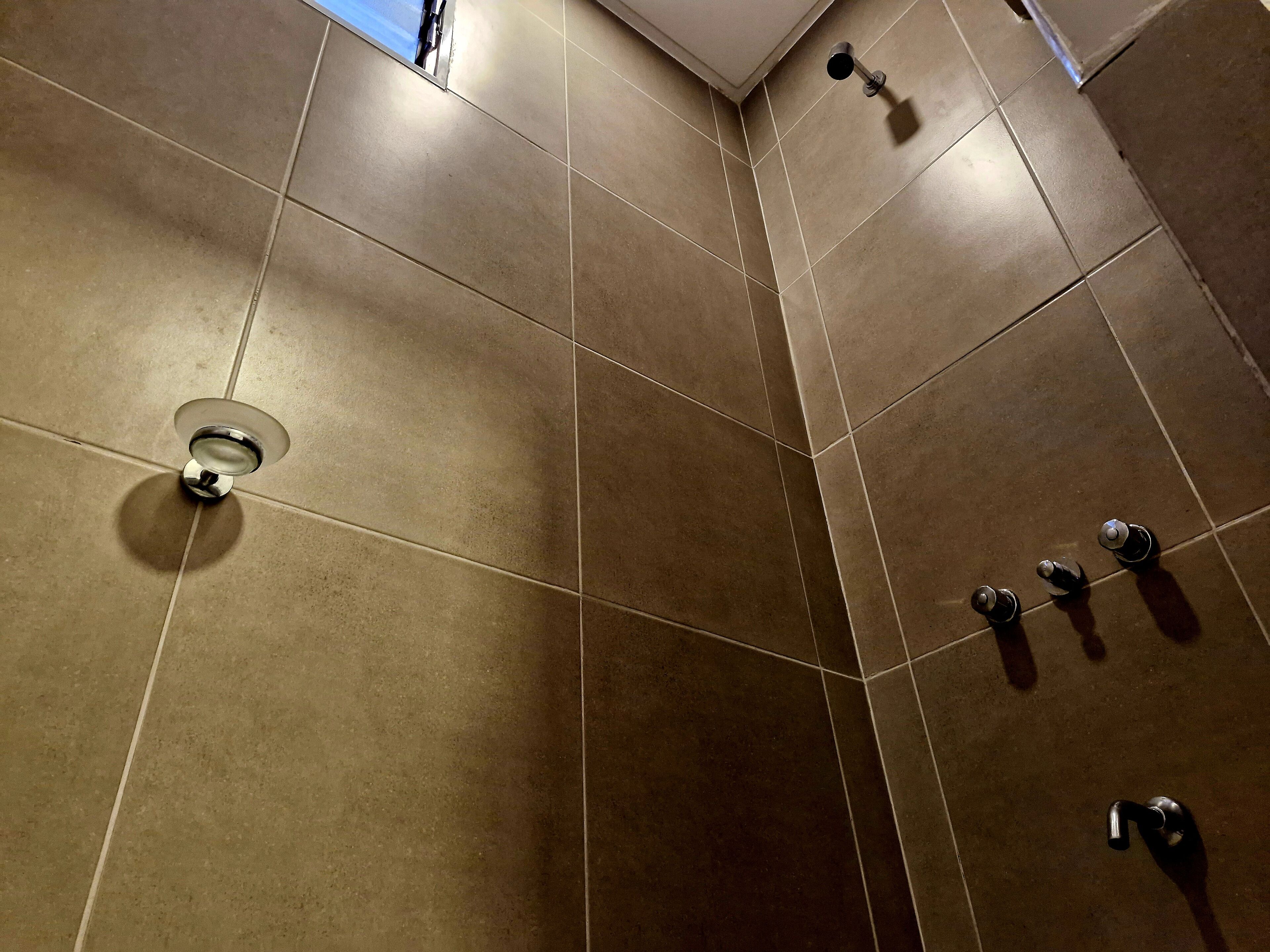 Un baño moderno con baldosas marrones grandes, una lámpara en el techo y tres grifos fijos en la pared.