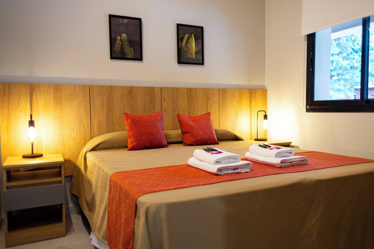 Une chambre d’hôtel avec une tête de lit en bois, deux oreillers rouges, un lit recouvert d’une couverture rouge, deux tables de chevet avec des lampes et deux tableaux encadrés accrochés au mur.