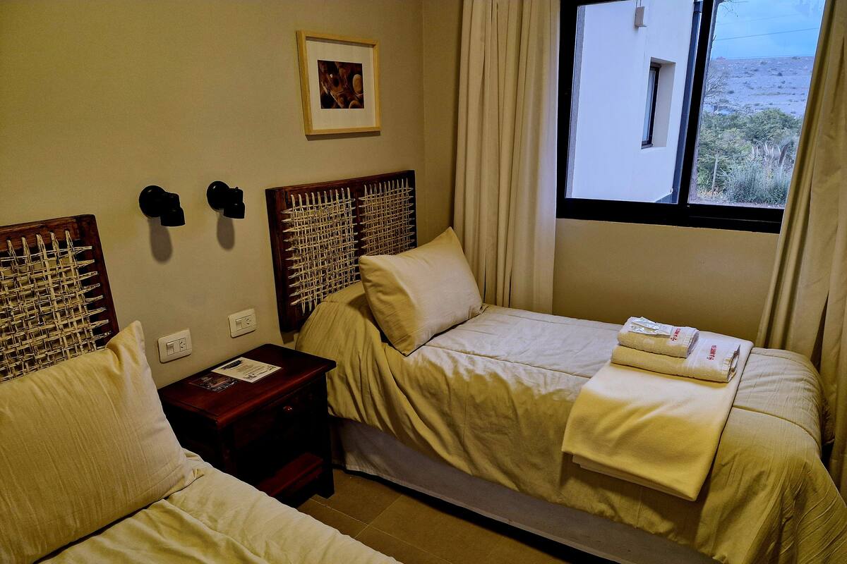 Une chambre d’hôtel avec deux lits, une tête de lit en bois, une table de chevet avec une lampe, une fenêtre avec des rideaux et un tableau encadré au mur.