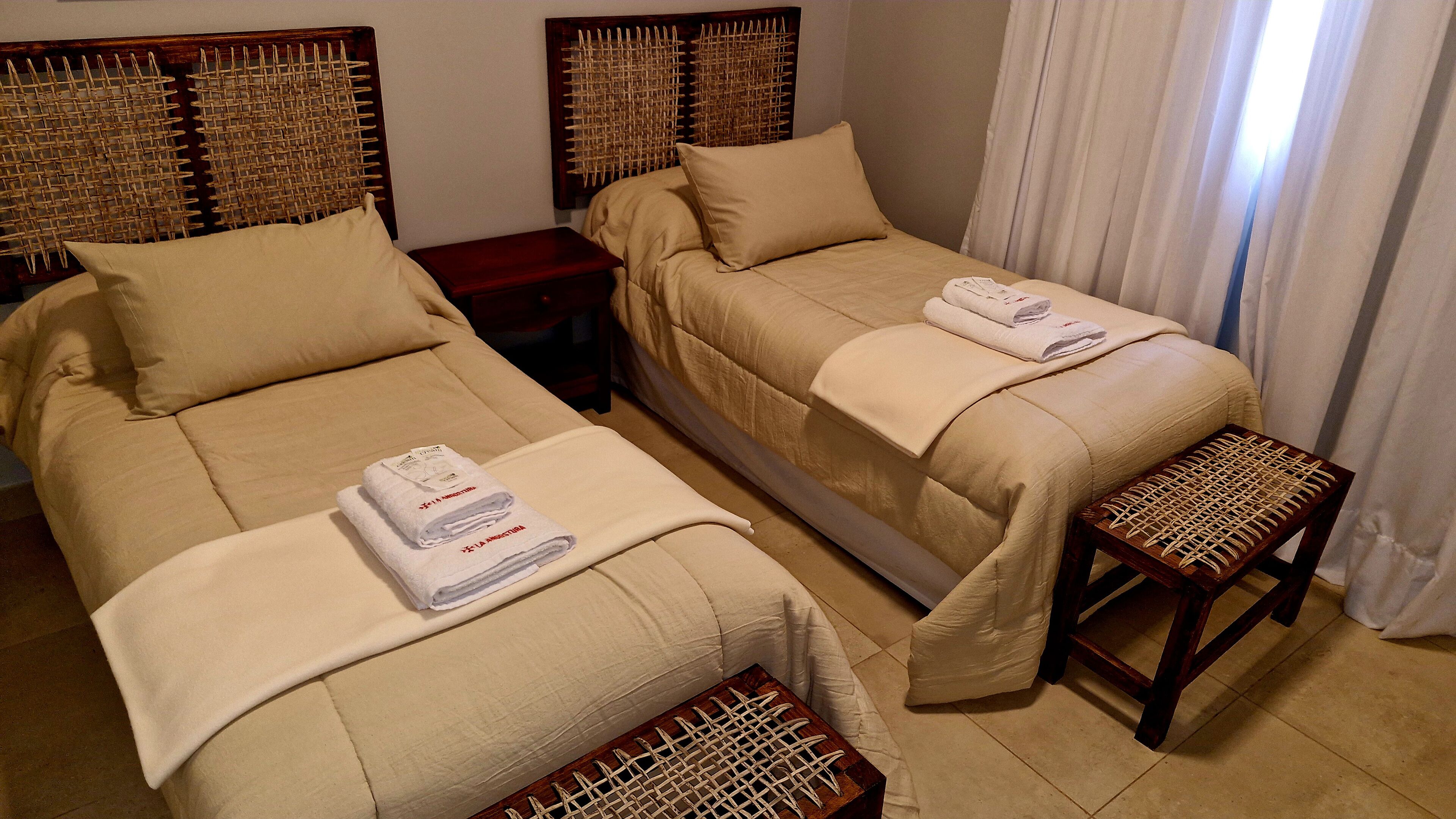A hotel room with two single beds, each with a wooden headboard, a small wooden bench, and a pair of folded towels on the beds.