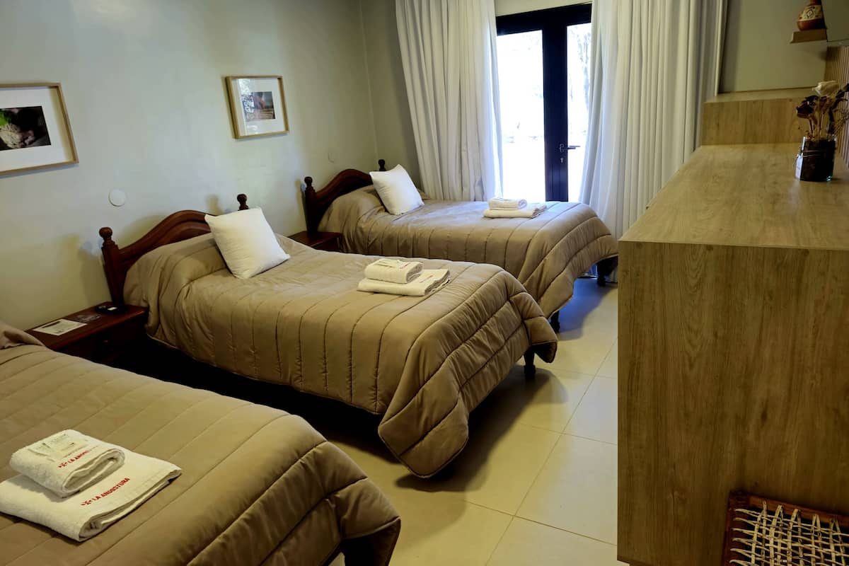 Une chambre d’hôtel avec trois lits, chacun recouvert d’une couette beige et agrémenté d’oreillers blancs. On y trouve une table de chevet en bois avec une lampe, ainsi qu’un tableau encadré au mur. Un bureau en bois avec un vase de fleurs séchées est également visible.