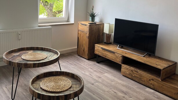 En moderne stue med et stort vindue, to runde træborde med flettede underlayere, et tv-bord i træ og hængende pendellamper.