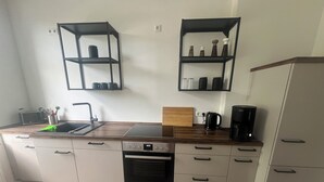 Et kompakt køkken med opvaskemaskine, ovn og kaffemaskine. To sorte hylder på væggen, oven over oven, med diverse køkkenartikler.