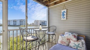 A balcony with a view of buildings across a body of water, a small round table with a candle, and metal chairs. - Peaceful, 2 BD, 2 BA Condo w/ Water Views, Pool and Free Activities Including Golf! (Ocean View)