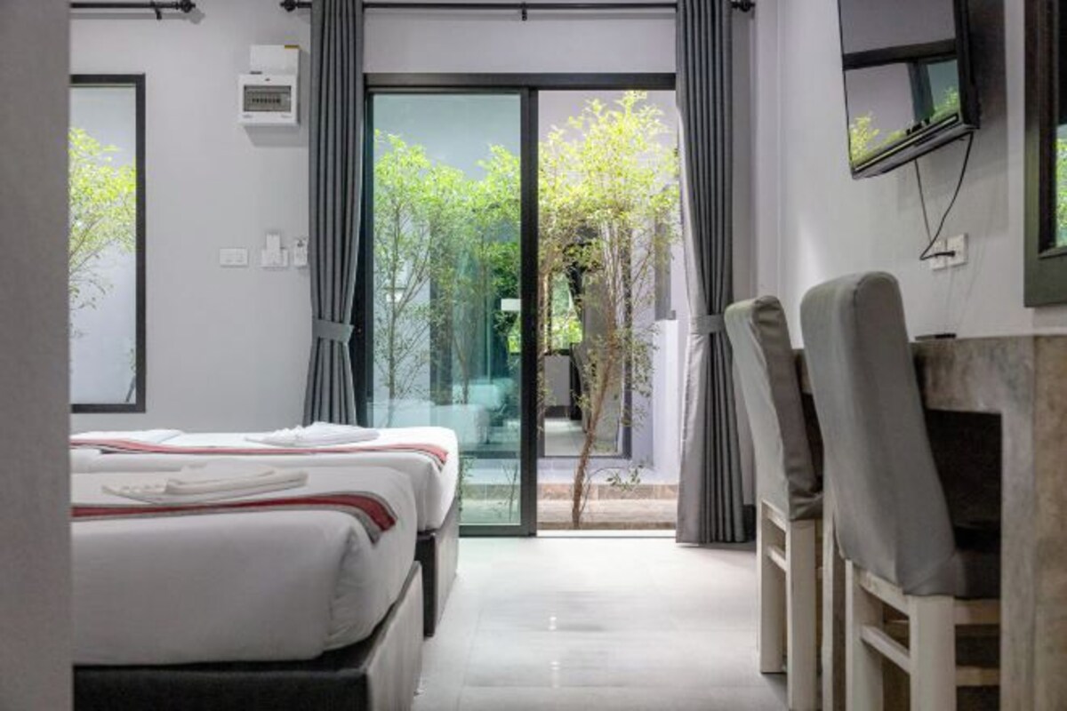 A modern hotel room with a bed, a desk with chairs, a TV, and a sliding glass door leading to an outdoor area with greenery.