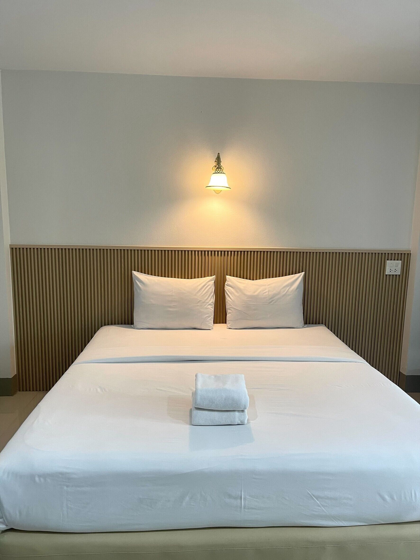 Habitación de hotel con una cama grande, dos almohadas, una lámpara de noche y una toalla doblada sobre la cama.